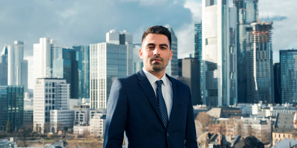 Der Fachanwalt für Arbeitsrecht Vassilios Mpouras ist im Anzug auf dem Dach seiner Kanzlei vor der Skyline von Frankfurt zu sehen.